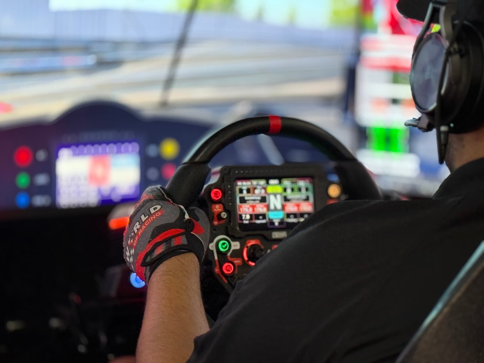 A driver in a headset gripping the wheel of a World of Racing sim, the track on the screens ahead